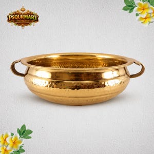 May include: A polished brass bowl featuring a hammered texture on the exterior, a wide rim, and two curved side handles. The bowl has a rounded base and a smooth interior surface. The logo for PSQURMART ONLINE SHOP is visible in the top left corner.
