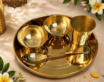 Op de afbeelding: Een gouden puja thali set van messing, inclusief een grote ronde plaat, twee kleine kommen, een beker en een lepel. De set is gerangschikt op een beige oppervlak, met witte en gele bloemen op de achtergrond. Het logo "PSQURMART ONLINE SHOP" is zichtbaar.