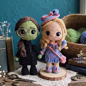 May include: Two amigurumi dolls made of yarn. One doll has green skin, brown hair, and round wire glasses, wearing a black dress. The other doll has blonde hair, a blue hat with a pink bow, and a blue dress with pink accents, holding a pink bag.