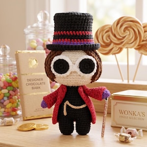 May include: A crocheted doll resembling a character from a children's book, wearing a black top hat, red coat, and holding a cane. The doll has large white-rimmed glasses and is surrounded by candy and chocolate bars.