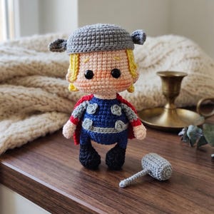 Può includere: Una bambola amigurumi all'uncinetto di un personaggio con capelli biondi, che indossa un elmo grigio con ali laterali, una tuta blu con accenti circolari grigi e un mantello rosso. La bambola ha occhi a perla neri e un martello grigio.