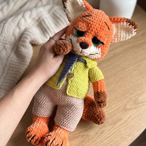Puede incluir: Muñeco de zorro de ganchillo hecho a mano con pelaje naranja, marrón y crema. El zorro lleva una chaqueta amarilla, pantalones cortos color beige y una corbata azul y gris. El zorro tiene ojos verdes y una nariz marrón. La muñeca está sobre una superficie de madera.