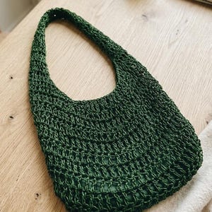 Op de afbeelding: Een donkergroene, geweven schoudertas met een afgeronde vorm en een enkele band. De tas heeft een textuur, open geweven ontwerp en een glad, effen groene binnenkant. De tas ligt op een lichtgekleurd houten oppervlak.