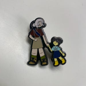 May include: Enamel pin featuring a cartoon illustration of two figures, an adult and a child, holding hands. The adult figure wears a red top, tan skirt, and green boots. The child wears a blue dress and yellow shoes. The pin has a silver outline.