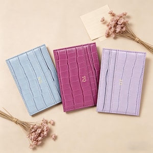 Könnte beinhalten: Drei Leder-Kartenetuis in Pastellfarben: Hellblau, Magenta und Lavendel. Jedes Kartenetui hat eine Krokodiltextur und goldfarbene Initialen. Die Kartenetuis sind auf einer beigen Oberfläche mit Trockenblumen und einem kleinen Notizblock angeordnet.