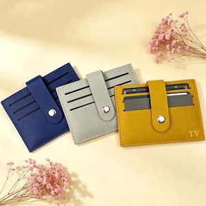 Puede incluir: Tres tarjeteros rectangulares en azul marino, gris claro y amarillo mostaza. Cada uno cuenta con una correa vertical central con un cierre de botón a presión de metal plateado, varias ranuras horizontales para tarjetas y las letras TY en el amarillo.