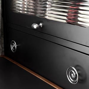 May include: Close-up of a black cabinet with silver hardware. The cabinet has two drawers with spiral-shaped pulls and a glass-fronted upper section. The interior of the cabinet is visible through the glass. A patterned wallpaper is visible on the left.