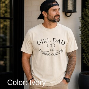 May include: Ivory-colored t-shirt with the text "GIRL DAD RAISING QUEENS" in black. The design includes a heart symbol. The shirt is short-sleeved and has a crew neck.
