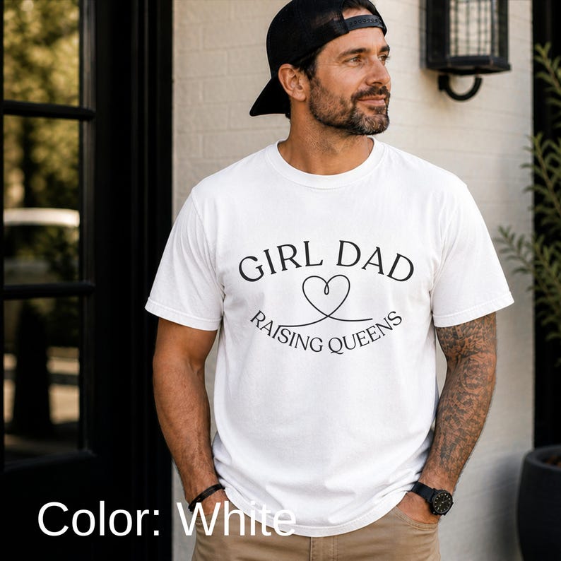 May include: White t-shirt with the text "GIRL DAD RAISING QUEENS" in black. The design includes a heart symbol. The shirt is short-sleeved.