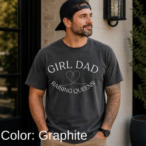 May include: Graphite-colored t-shirt with the text "GIRL DAD RAISING QUEENS" in white. The design includes a heart symbol. The shirt is worn with a black cap and watch.