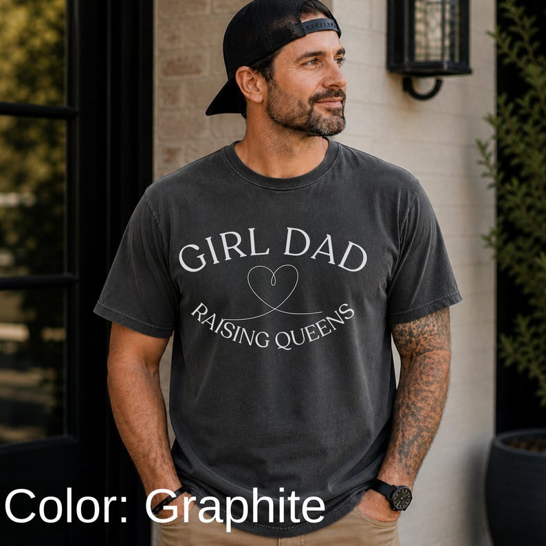 May include: Graphite-colored t-shirt with the text "GIRL DAD RAISING QUEENS" in white. The design includes a heart symbol. The shirt is worn with a black cap and watch.