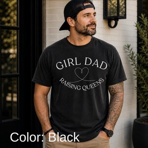 May include: Black t-shirt with the text "GIRL DAD RAISING QUEENS" in white. The design includes a heart symbol. The shirt is worn by a person wearing a black cap and watch.