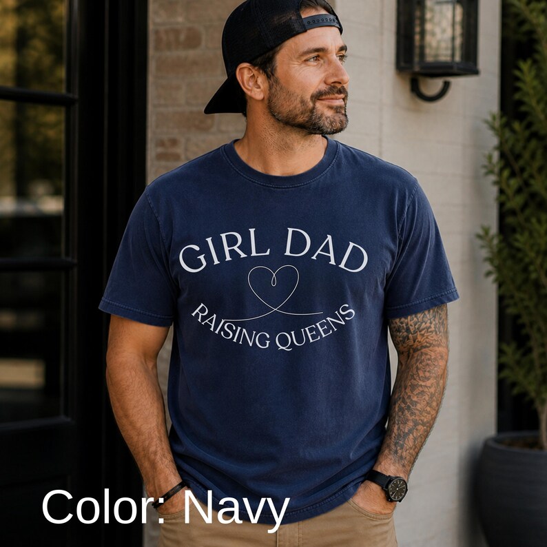 May include: Navy blue t-shirt with the text "GIRL DAD RAISING QUEENS" in white script and a heart graphic. The shirt is a crew neck style and is made of a soft material. The color is navy.