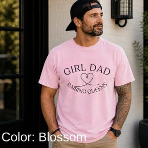 May include: A light pink t-shirt with the text "GIRL DAD RAISING QUEENS" in black. The design includes a heart symbol. The shirt is short-sleeved and appears to be made of a soft material.