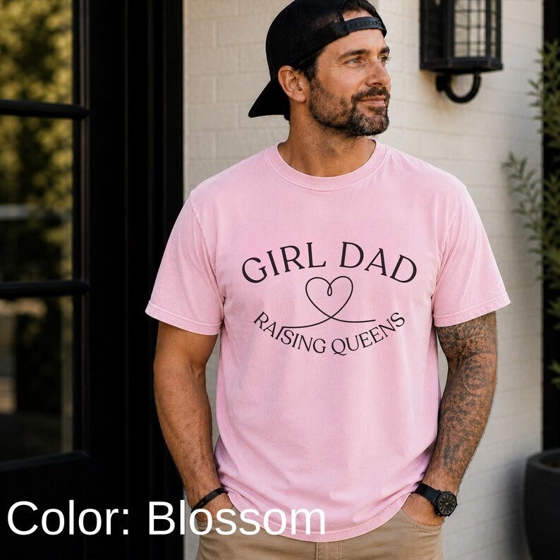 May include: A light pink t-shirt with the text "GIRL DAD RAISING QUEENS" in black. The design includes a heart symbol. The shirt is short-sleeved and appears to be made of a soft material.