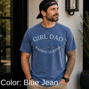 May include: Blue jean colored t-shirt with the text "GIRL DAD RAISING QUEENS" in white. The design includes a heart shape. The shirt has short sleeves and a crew neck. The color is described as blue jean.