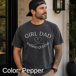 May include: Pepper-colored t-shirt with the text "GIRL DAD RAISING QUEENS" in white. The design includes a heart symbol. The shirt is short-sleeved and has a vintage, worn look.