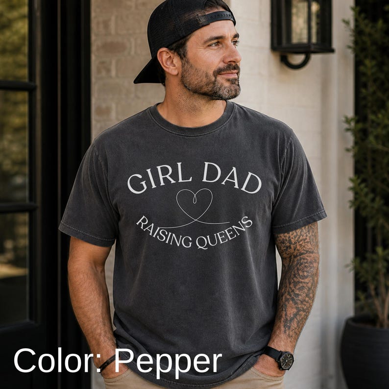 May include: Pepper-colored t-shirt with the text "GIRL DAD RAISING QUEENS" in white. The design includes a heart symbol. The shirt is short-sleeved and has a vintage, worn look.