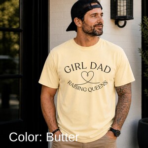 May include: Butter-colored t-shirt with the text "GIRL DAD RAISING QUEENS" in black script. The design includes a heart symbol. The shirt is short-sleeved and appears to be made of a soft material.