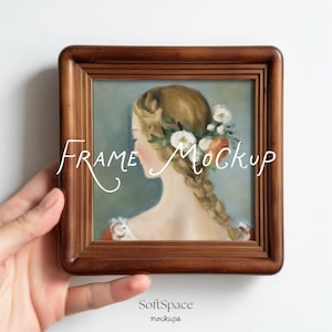 Könnte beinhalten: Ein quadratischer Holzbilderrahmen mit einem Gemälde einer Frau mit Zopf und Blumen im Haar. Die Worte "Frame Mockup" sind in Schreibschrift über das Bild geschrieben. Der Rahmen wird in einer Hand gehalten.