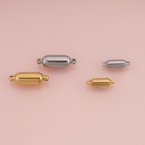 May include: Four cylindrical jewelry clasps in two sizes and two finishes. Two clasps are gold-toned and two are silver-toned. Each clasp features a smooth, rounded barrel shape with a small circular loop extending from each end for attachment.