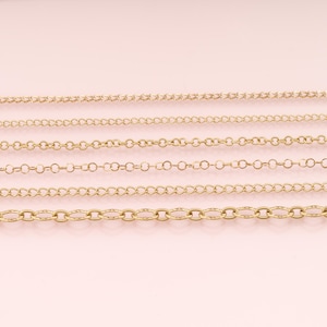 May include: Six distinct gold-toned metal chains arranged horizontally. The collection includes various link styles, ranging from fine, delicate cable chains to a thicker, textured oval link chain at the bottom, all featuring a polished finish.