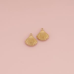Op de afbeelding: Twee goudkleurige metalen schelpbedels, elk met een getextureerd, waaiervormig ontwerp en een kleine ronde lus aan de bovenkant voor bevestiging. De bedels hebben een metallic afwerking en een gedetailleerd, verhoogd patroon van schelpribbels.