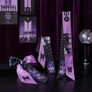 May include: A display of BTS merchandise, including purple and black scarves, posters, and a light stick. The scarves feature the BTS logo and geometric patterns. Posters display the BTS logo and silhouettes of the band members. The light stick has the word "ARMY" on it.