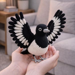 May include: A handmade crochet magpie bird with outstretched wings. The bird features a black body, white accents on the wings, and a white chest. The magpie is held in open hands, showcasing the craft's detail and the bird's lifelike appearance.