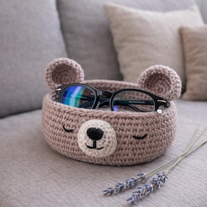 May include: A handmade, light brown crochet basket shaped like a bear's face, complete with ears, eyes, and a nose. The basket holds a pair of black-framed glasses. A sprig of lavender rests beside the basket on a gray surface.