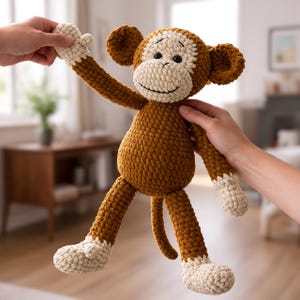 Könnte beinhalten: Eine handgefertigte Häkelpuppe in Form eines Affen, in Braun- und Cremetönen. Das Stofftier hat ein lächelndes Gesicht, lange Arme und Beine sowie einen eingerollten Schwanz. Der Affe wird von zwei Händen gehalten.