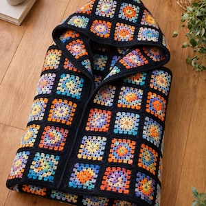 May include: A black crochet hooded vest with a colorful granny square pattern. The squares feature vibrant hues of orange, blue, yellow, and purple. The vest is displayed on a wooden surface.