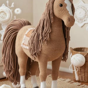 Op de afbeelding: Een handgemaakt gehaakt paardenspeelgoed in bruin en beige. Het paard heeft een lange, vloeiende manen en staart, met witte markeringen op het gezicht en de benen. Het staat op een houten voet, met een gedetailleerd, getextureerd ontwerp. Een charmant decoratief stuk.