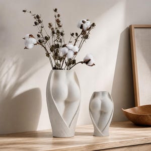 Könnte beinhalten: Zwei cremeweiße Keramikvasen mit einem skulpturalen menschlichen Torso-Design. Die größere Vase enthält einen Strauß aus getrockneten braunen Stielen und weißen Baumwollblüten. Die Vasen haben glatte, matte Oberflächen und geschwungene Konturen.