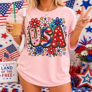 May include: A light pink t-shirt with a colorful "USA" graphic. The letters are decorated with red, white, and blue patterns and surrounded by flowers and stars. The shirt is a casual style, perfect for patriotic events.