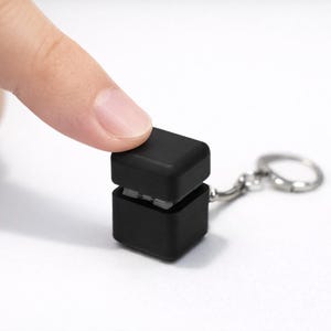 Könnte beinhalten: Ein kleines, schwarzes, würfelförmiges Schlüsselanhänger-Zubehör. Ein Finger drückt auf den oberen Teil. Der Schlüsselanhänger hat einen silberfarbenen Ring. Der Hintergrund ist weiß.