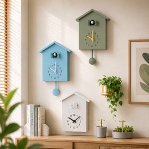 May include: Three house-shaped wall clocks in light blue, sage green, and white. Each clock features a small door at the top, a circular clock face with numbers, and a pendulum. The blue and green clocks have visible pendulums hanging below.