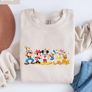 May include: A sand-coloured crewneck sweatshirt featuring a colourful graphic print of Goofy, Minnie Mouse, Mickey Mouse, Donald Duck, Daisy Duck, and Pluto holding various desserts, with the word SAND printed in the top left corner of the image.