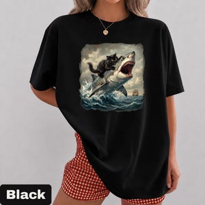 May include: Black t-shirt featuring a graphic of a black cat riding a shark through ocean waves. The shark has its mouth open, and a ship is visible in the background. The word "Black" is printed at the bottom.