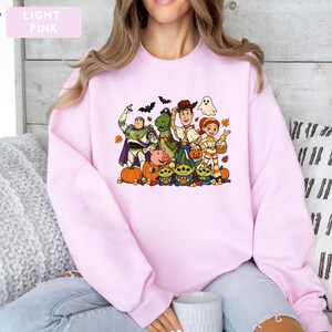 Puede incluir: Una sudadera de cuello redondo rosa claro con un estampado gr&aacute;fico colorido de varios personajes animados vestidos con disfraces de Halloween, incluyendo calabazas, un fantasma y murci&eacute;lagos, con el texto LIGHT PINK en la esquina superior izquierda.