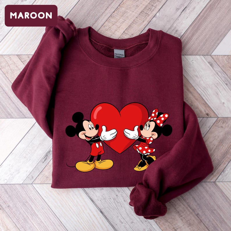 May include: A maroon long-sleeve crewneck sweatshirt featuring a graphic print of Mickey and Minnie Mouse holding a large red heart between them. The text MAROON is visible in the upper left corner. The sweatshirt has a ribbed collar and cuffs.