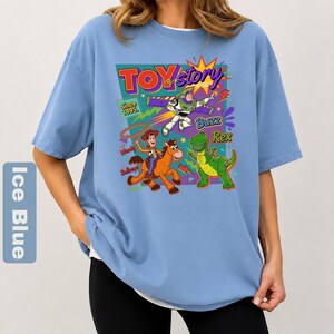 May include: Ice blue t-shirt featuring a colorful graphic of Toy Story characters, including Woody, Buzz Lightyear, and Rex. The design includes the text "TOY STORY" and "Since 1995". The shirt has short sleeves and a crew neck.