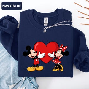 May include: A navy blue crewneck sweatshirt featuring a graphic of Mickey and Minnie Mouse holding a large red heart. The sweatshirt has long sleeves and a ribbed collar. A silver chain necklace with a small circular pendant rests on the neckline.