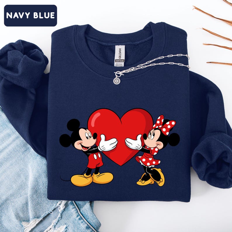 May include: A navy blue crewneck sweatshirt featuring a graphic of Mickey and Minnie Mouse holding a large red heart. The sweatshirt has long sleeves and a ribbed collar. A silver chain necklace with a small circular pendant rests on the neckline.