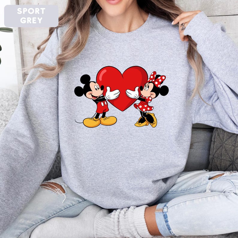 May include: A sport grey crewneck sweatshirt featuring a graphic print of Mickey and Minnie Mouse holding a large red heart between them. The design shows Mickey in red shorts and yellow shoes, and Minnie in a red polka dot dress and bow.