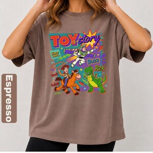 May include: Espresso-colored t-shirt featuring a colorful Toy Story graphic. The design includes Woody, Buzz Lightyear, and Rex, with the words "Toy Story" and "Since 1995." The shirt has a relaxed fit and short sleeves.