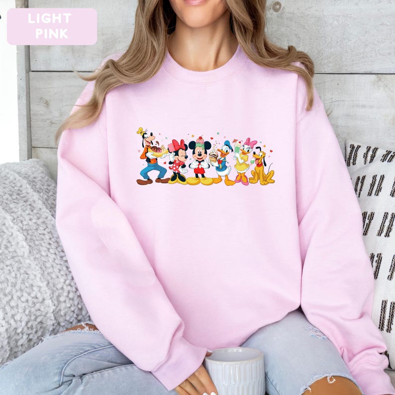 May include: A light pink crewneck sweatshirt featuring a colourful graphic print of Disney characters including Goofy, Minnie Mouse, Mickey Mouse, Donald Duck, Daisy Duck, and Pluto, all arranged in a line with small red hearts scattered around them.