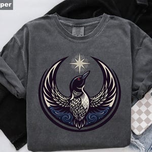 May include: Pepper-colored t-shirt featuring a graphic of a bird with outstretched wings, a crescent moon, and a star. The bird has black and white feathers and red eyes. The design is centered on the shirt.
