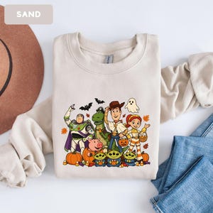 Puede incluir: Una sudadera de cuello redondo color arena con un estampado gr&aacute;fico de personajes de Toy Story disfrazados para Halloween. El dise&ntilde;o incluye a Buzz Lightyear, Rex, Woody, Jessie, Hamm y tres alien&iacute;genas rodeados de calabazas, murci&eacute;lagos y un fantasma.