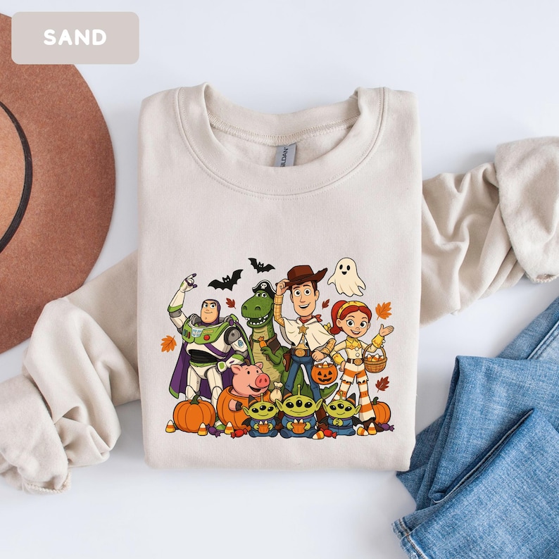 Puede incluir: Una sudadera de cuello redondo color arena con un estampado gr&aacute;fico de personajes de Toy Story disfrazados para Halloween. El dise&ntilde;o incluye a Buzz Lightyear, Rex, Woody, Jessie, Hamm y tres alien&iacute;genas rodeados de calabazas, murci&eacute;lagos y un fantasma.